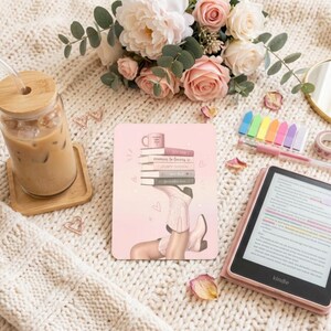 May include: A rectangular e-reader skin featuring a pink gradient background with an illustration of a stack of books balanced on a pair of light-colored cowboy boots. The design includes small heart accents and a coffee mug graphic on the top book.