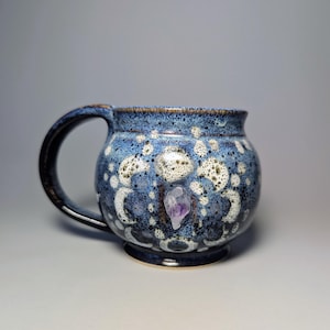 May include: A handmade ceramic mug with a blue and white speckled glaze. The mug features a dark handle and a small, purple amethyst crystal embedded in the front. The mug is designed for beverages.