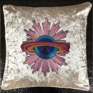 May include: A cream-colored velvet pillow with a pink and blue embroidered design of a planet with rings. The planet is surrounded by a pink flower-like design.