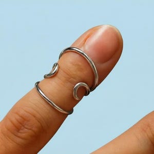 May include: A silver-colored finger ring with a unique, open design. The ring wraps around the finger with curved accents, creating an elegant and modern look. The ring is shown against a light blue background.