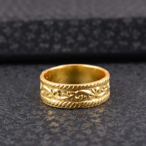 May include: A gold ring with an embossed design. The ring has a repeating pattern of swirling lines and rope-like borders. The ring is set against a dark, textured background, highlighting its detailed craftsmanship.