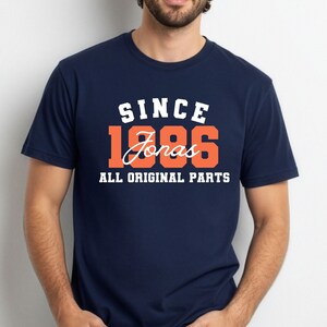 May include: A navy blue t-shirt featuring the text "SINCE 1986 Jonas ALL ORIGINAL PARTS" in white and orange. The shirt has a crew neck and is likely made from a soft fabric.