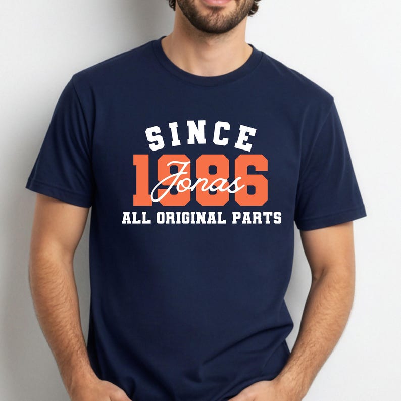 May include: A navy blue t-shirt featuring the text "SINCE 1986 Jonas ALL ORIGINAL PARTS" in white and orange. The shirt has a crew neck and is likely made from a soft fabric.