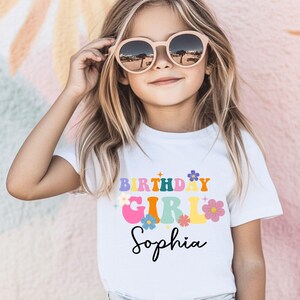 Pode incluir: Uma camiseta branca com as palavras "BIRTHDAY GIRL Sophia" em letras coloridas em estilo retr&ocirc;. A camiseta apresenta detalhes florais e &eacute; usada por uma menina com &oacute;culos de sol redondos.