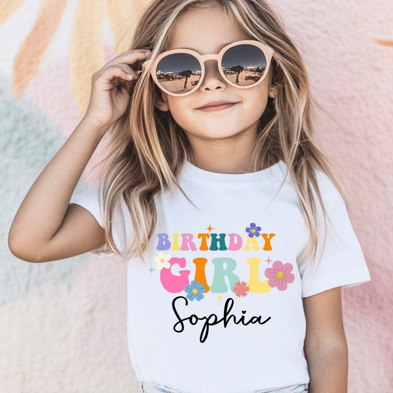 Pode incluir: Uma camiseta branca com as palavras "BIRTHDAY GIRL Sophia" em letras coloridas em estilo retr&ocirc;. A camiseta apresenta detalhes florais e &eacute; usada por uma menina com &oacute;culos de sol redondos.