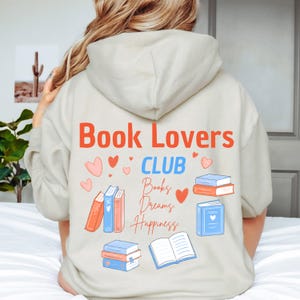 Könnte beinhalten: Ein beigefarbener Kapuzenpullover mit einem Aufdruck auf der Rückseite, der den Text "Book Lovers CLUB" in Orange und Blau zeigt. Darunter stehen die Wörter "Books, Dreams, Happiness" in Schreibschrift, umgeben von Buch- und Herzmotiven.