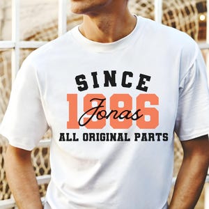 May include: White t-shirt with the text "SINCE 1986 Jonas ALL ORIGINAL PARTS" in black and coral lettering. The shirt has a crew neck and short sleeves.