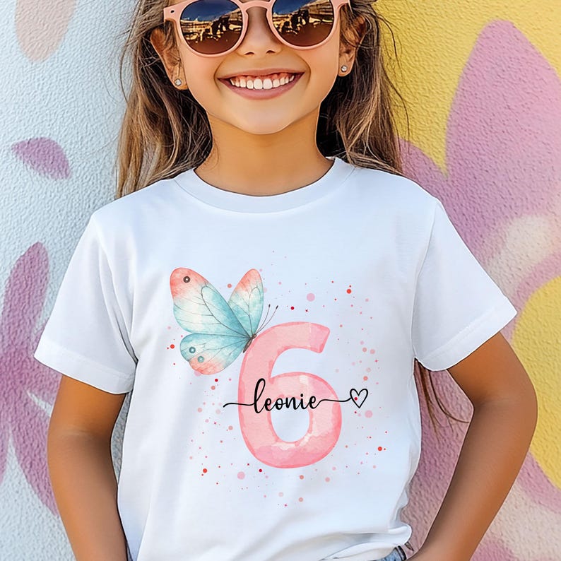 Op de afbeelding: Wit T-shirt met een aquarelontwerp. Het heeft het roze nummer 6, een blauwe en roze vlinder, de naam "leonie" in script en een hart. Het ontwerp heeft roze aquarel spikkels.