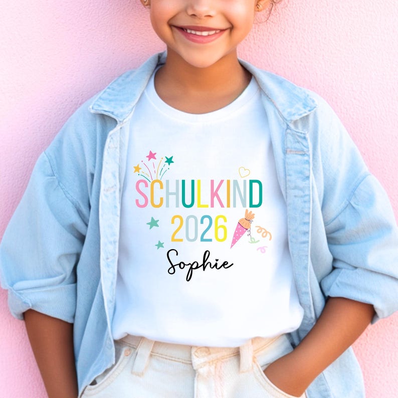 May include: A white t-shirt featuring the colorful text SCHULKIND 2026 and the name Sophie in black script. The design includes multi-colored stars, a small heart, and a pink polka-dot cone graphic with ribbons, layered under a light blue denim shirt.