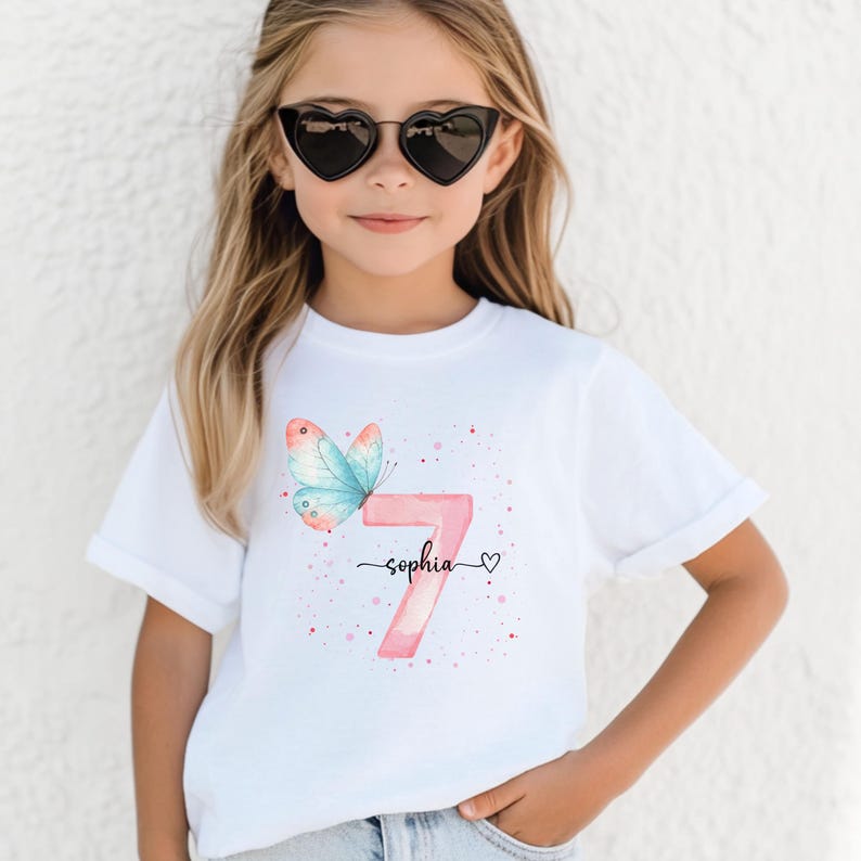May include: White t-shirt with a watercolour design. It features a pink number 7, a blue and pink butterfly, and the name "Sophia" in script. The design includes pink speckles and a small heart.