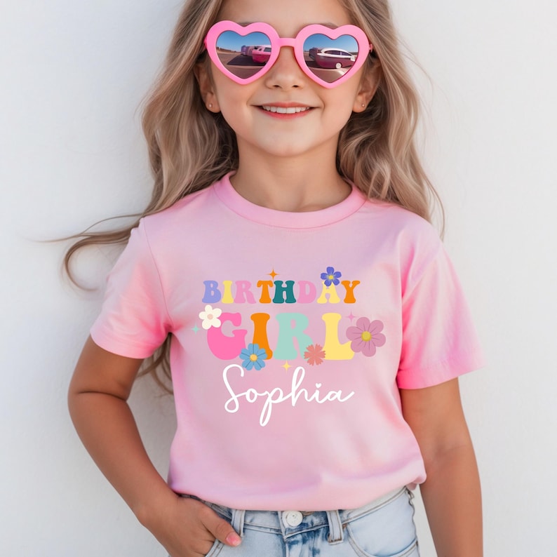 Pode incluir: Uma camiseta rosa com as palavras "BIRTHAY GIRL Sophia" em letras coloridas em estilo retr&ocirc; e detalhes florais. A menina est&aacute; usando &oacute;culos de sol em forma de cora&ccedil;&atilde;o. A camisa &eacute; de manga curta.