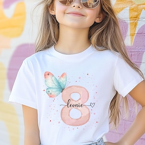 Op de afbeelding: Wit T-shirt met een aquarelontwerp. Het heeft het roze nummer 8, een vlinder en de naam "Leonie" in cursief met een hartsymbool. Kleine rode stippen maken het ontwerp compleet. Kinderverjaardagsthema.
