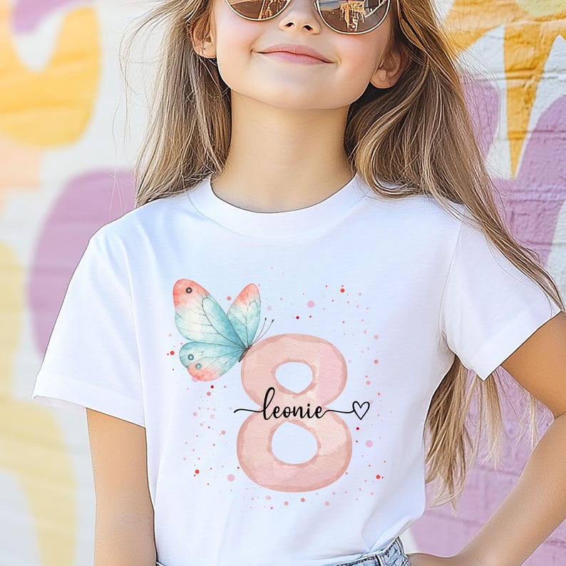 Op de afbeelding: Wit T-shirt met een aquarelontwerp. Het heeft het roze nummer 8, een vlinder en de naam "Leonie" in cursief met een hartsymbool. Kleine rode stippen maken het ontwerp compleet. Kinderverjaardagsthema.