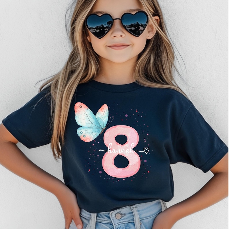May include: Navy blue t-shirt with a watercolour butterfly and the number 8 in pink. The name "Hannah" is written in cursive below the number, with a small heart. A birthday-themed graphic tee for children.
