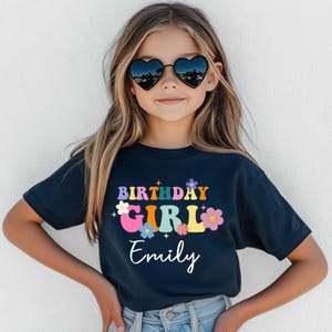 Pode incluir: Uma camiseta azul marinho com as palavras "BIRTHAY GIRL Emily" em letras coloridas em estilo retr&ocirc;. O design inclui flores e uma fonte groovy. A menina est&aacute; usando &oacute;culos de sol em forma de cora&ccedil;&atilde;o.