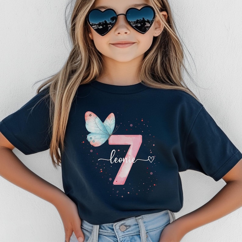 Op de afbeelding: Marineblauw T-shirt met een roze nummer 7, een vlinder en de naam "Leonie". De vlinder heeft blauwe en roze vleugels. Het ontwerp bevat kleine roze stippen.
