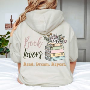 Könnte beinhalten: Ein beigefarbener Kapuzenpullover mit einer Grafik auf der Rückseite, die einen Stapel aus fünf Büchern mit Blumen darauf zeigt. Der Text lautet Book lovers in rosa und dunkelgrüner Schrift, darunter Read. Dream. Repeat. in orange.