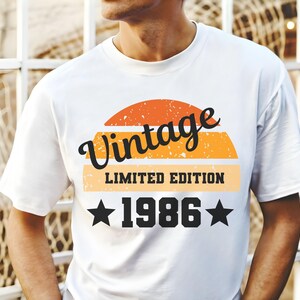 Op de afbeelding: Wit t-shirt met een retro design. Het ontwerp bevat het woord "Vintage" in zwart script boven een zonsondergangafbeelding. Daaronder staan de woorden "LIMITED EDITION" en het jaar "1986" met steraccenten.