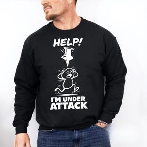 Puede incluir: Sudadera negra con las palabras "HELP!" y "I'M UNDER ATTACK" en blanco. Se representa una figura de dibujos animados con una expresión de pánico, con una flecha blanca que apunta hacia abajo.