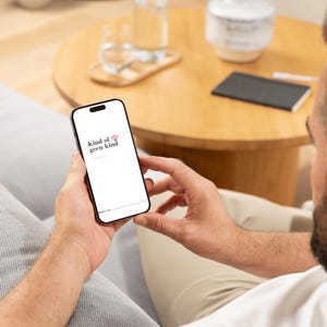 May include: A black smartphone displaying the text "kind of geen kind" in a white font with a pink flower graphic. The phone is held in a person's hands, with a blurred background of a wooden table and sofa.