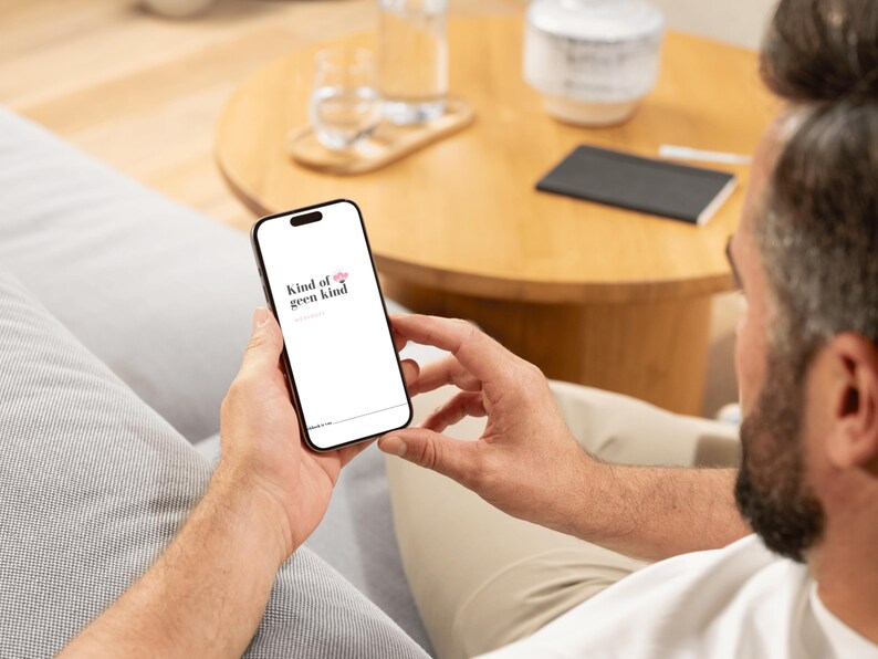 May include: A black smartphone displaying the text "kind of geen kind" in a white font with a pink flower graphic. The phone is held in a person's hands, with a blurred background of a wooden table and sofa.