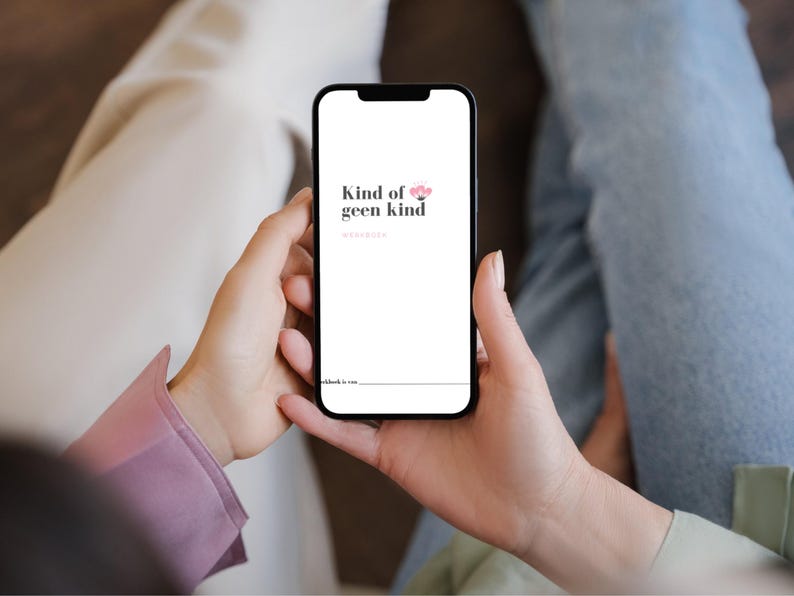 May include: A smartphone displays the text "Kind of geen kind" in black, with a pink flower graphic. The phone is held by a person, with a pink sleeve visible. The background shows light-colored pants and blue jeans.