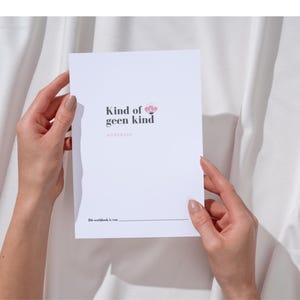 May include: A white workbook with the text "Kind of geen kind" and a floral design is held by two hands. The workbook's title is in a dark font, with the word "workbook" below. The background is a white fabric.
