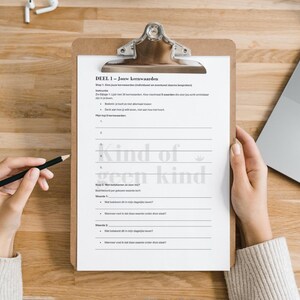 May include: A wooden clipboard with a white paper sheet featuring Dutch text and the phrase "kind of geen kind." A hand holds a pencil, poised to write. A laptop and earbuds are also visible on the wooden surface.