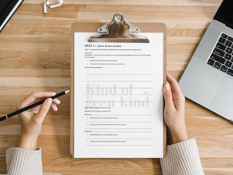 May include: A wooden clipboard with a white paper sheet featuring Dutch text and the phrase "kind of geen kind." A hand holds a pencil, poised to write. A laptop and earbuds are also visible on the wooden surface.