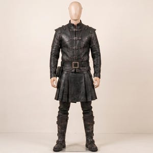 May include: A black leather outfit consisting of a long-sleeved jacket with multiple front buckles, a matching pleated kilt, a wide belt with a square buckle, and tall leather boots. The ensemble features structured shoulders and a dark finish.
