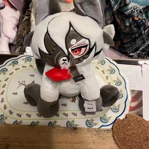 May include: A plush toy in the shape of a stylized animal with large ears, a white body, and gray accents. The toy has a detailed face with red eyes and a red bow tie. It sits on a floral patterned plate.