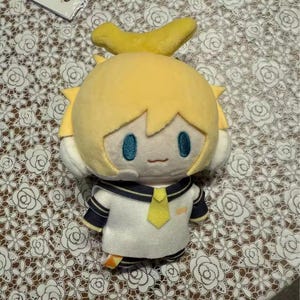 May include: A plush doll with yellow hair and a matching banana-shaped hair accessory. The doll has a white sailor-style uniform with navy blue accents and a yellow tie. The doll has blue eyes and a small smile.