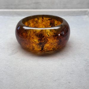 May include: A wide, round bracelet with a marbled amber and brown design. The bracelet has a glossy finish and a smooth, rounded shape. The interior of the bracelet has a swirling pattern of dark brown and amber colours.