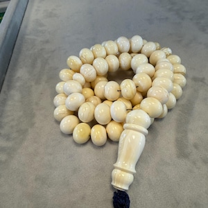 May include: Ivory-colored prayer beads with a decorative ivory-colored end piece and a dark blue tassel. The beads are round and appear to be made of a smooth material. The beads are arranged in a circular pattern.