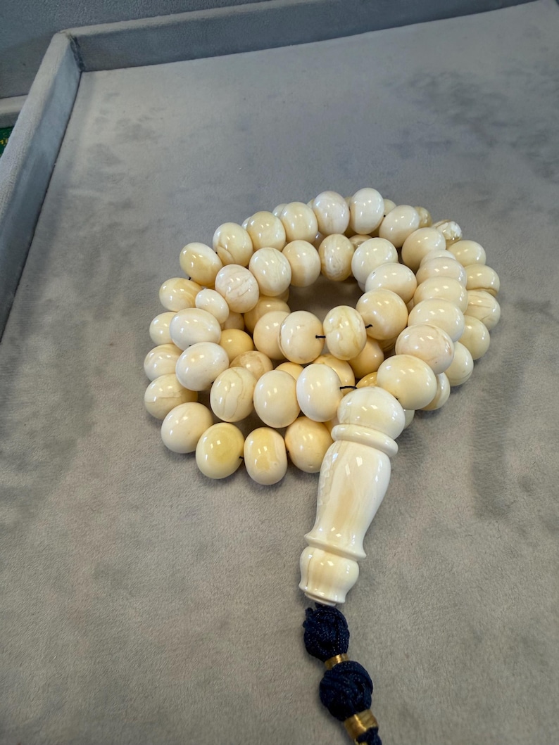 May include: Ivory-colored prayer beads with a decorative ivory-colored end piece and a dark blue tassel. The beads are round and appear to be made of a smooth material. The beads are arranged in a circular pattern.