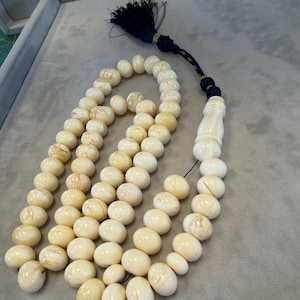 May include: A cream-colored prayer bead necklace with oval beads. The beads are strung together with a dark blue tassel and a decorative end piece. The beads have a marbled appearance and vary slightly in color.