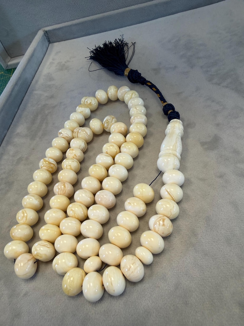 May include: A cream-colored prayer bead necklace with oval beads. The beads are strung together with a dark blue tassel and a decorative end piece. The beads have a marbled appearance and vary slightly in color.