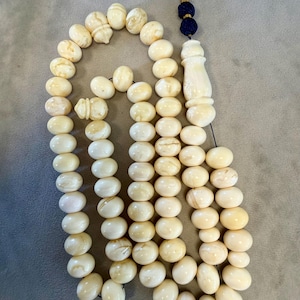 May include: Ivory-colored prayer beads with an ornate tassel. The beads are oval-shaped and strung together, with a decorative carved piece and a blue and gold tassel. The beads have a smooth, polished appearance.