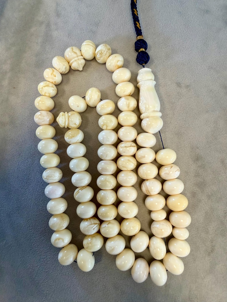 May include: Ivory-colored prayer beads with an ornate tassel. The beads are oval-shaped and strung together, with a decorative carved piece and a blue and gold tassel. The beads have a smooth, polished appearance.