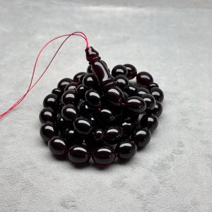 Puede incluir: Un juego de cuentas de oración de color burdeos oscuro, con una serie de cuentas lisas y redondas. Las cuentas están ensartadas con una borla burdeos a juego y un hilo rojo. Las cuentas están dispuestas sobre una superficie gris.