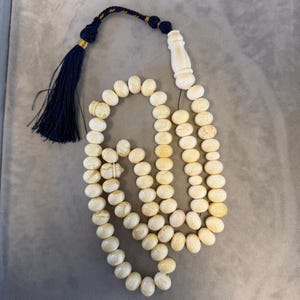 May include: Ivory-colored prayer beads with a navy blue tassel and decorative accents. The beads are oval-shaped and strung together, with a decorative ivory-colored piece at the top. The tassel is navy blue with gold and navy blue accents.