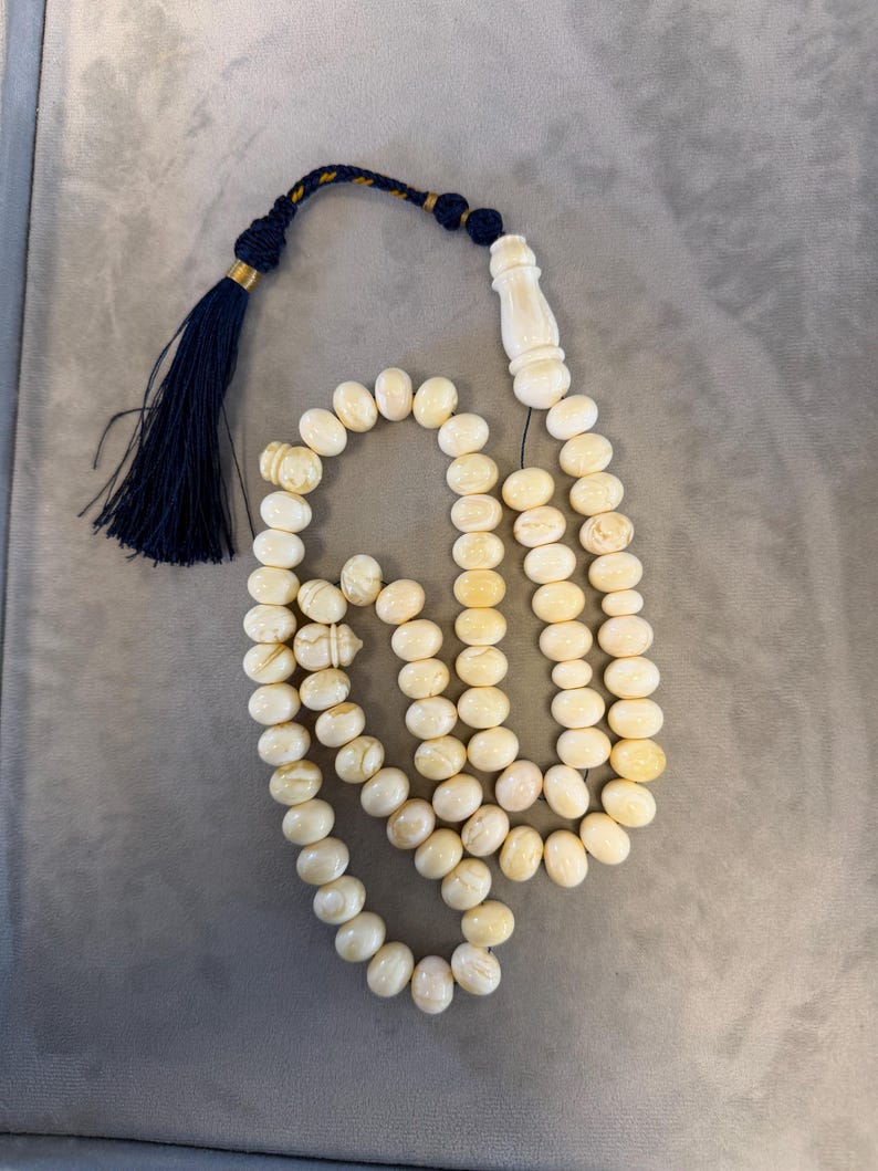 May include: Ivory-colored prayer beads with a navy blue tassel and decorative accents. The beads are oval-shaped and strung together, with a decorative ivory-colored piece at the top. The tassel is navy blue with gold and navy blue accents.