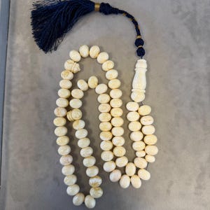 May include: Ivory-colored prayer beads with a tassel. The beads are oval-shaped and strung together. The tassel is dark blue with gold accents. The beads are likely made of a natural material.