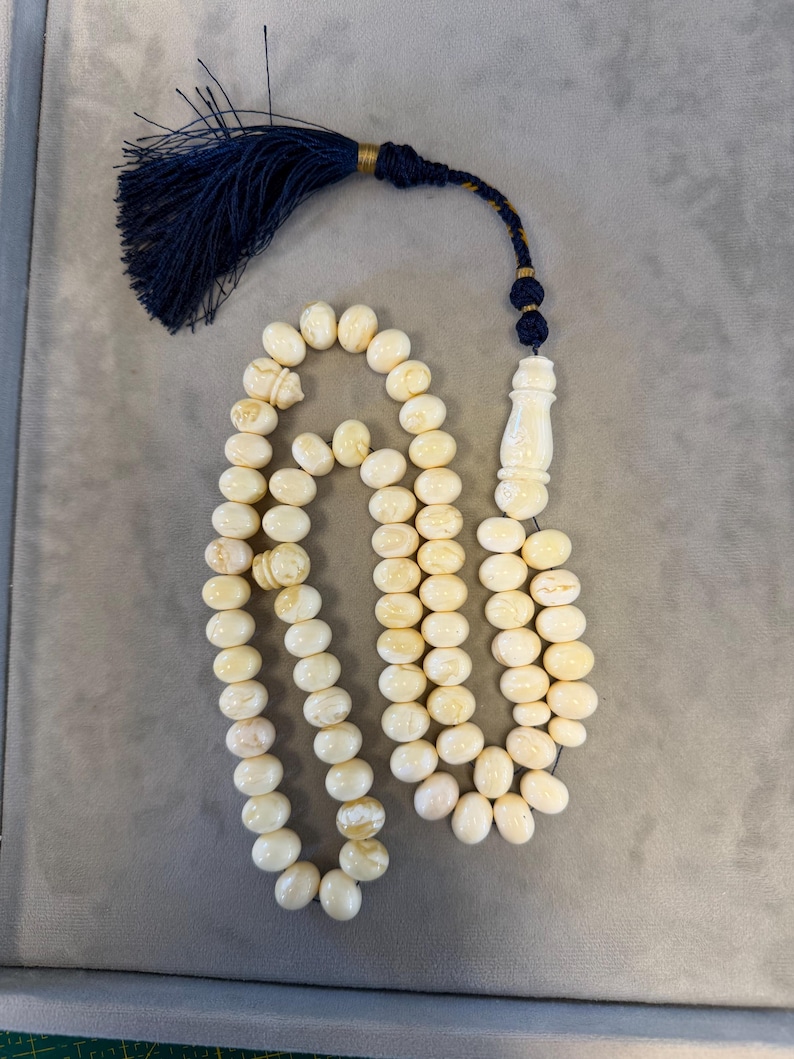 May include: Ivory-colored prayer beads with a tassel. The beads are oval-shaped and strung together. The tassel is dark blue with gold accents. The beads are likely made of a natural material.