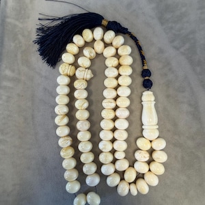 May include: Ivory-colored prayer beads with a navy blue tassel and decorative accents. The beads are oval-shaped and strung together, with a larger, elongated bead at the end. The tassel is made of navy blue threads.