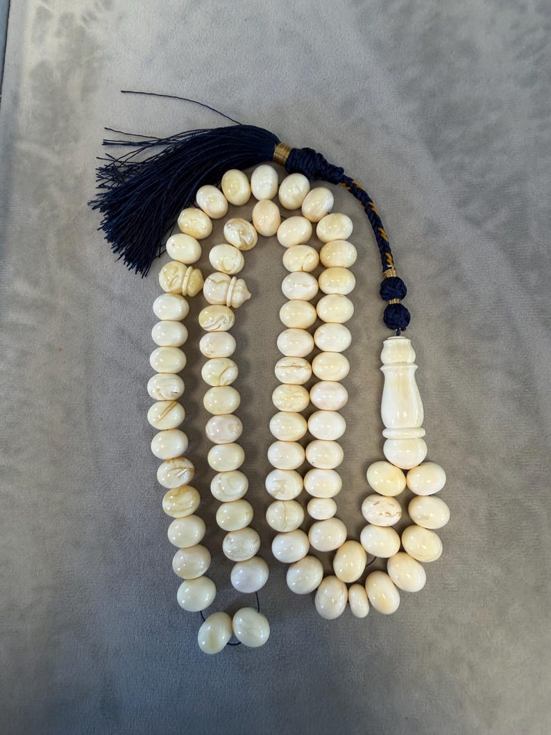 May include: Ivory-colored prayer beads with a navy blue tassel and decorative accents. The beads are oval-shaped and strung together, with a larger, elongated bead at the end. The tassel is made of navy blue threads.