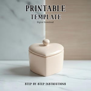 Puede incluir: Una caja de baratijas de cerámica rosa pálido en forma de corazón con tapa y pomo. El texto "PRINTABLE TEMPLATE" y "Digital Download" está en la parte superior. El texto "STEP BY STEP INSTRUCTIONS" está en la parte inferior. La caja está sobre mármol blanco.