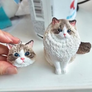 Puede incluir: Una detallada figura de un gato Ragdoll con ojos azules y un pelaje blanco y marrón esponjoso. Junto a ella, una cabeza de gato más pequeña, tamaño colgante, con un lazo plateado. Las figuras de gato están sobre una superficie blanca.