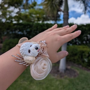 May include: A wrist corsage featuring a white dog figurine with a black nose and pink tongue, surrounded by beige fabric roses and sparkling rhinestones. The corsage is designed with a beige ribbon and pearl accents, perfect for a special occasion.