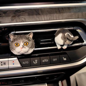 Könnte beinhalten: Zwei katzenförmige Lüftungsdekorationen für Autos. Eine ist ein Katzenkopf mit grauem und weißem Fell und gelben Augen. Die andere ist das Hinterteil einer Katze mit grauem und weißem Fell und rosa Pfoten. Die Dekorationen befinden sich an einer Autolüftung.
